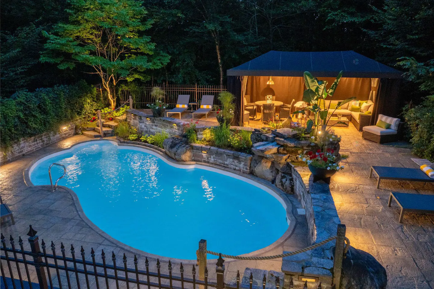 An aerial view of a backyard with a blue pool, stone patio, lounge chairs, and a covered gazebo with seating at dusk.