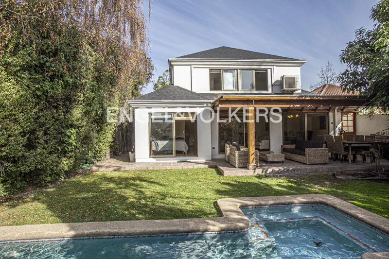 Backyard of a white two-story house with a pool, patio furniture under a wooden pergola, and lush green landscaping.