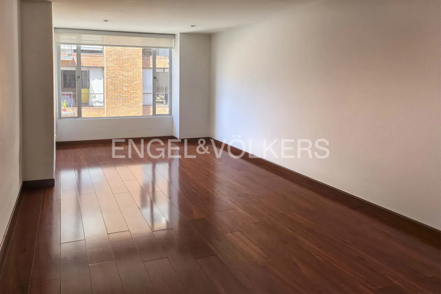Empty room with white walls, wood floors, and a large window showing a brick building. Engel & Völkers logo is visible.