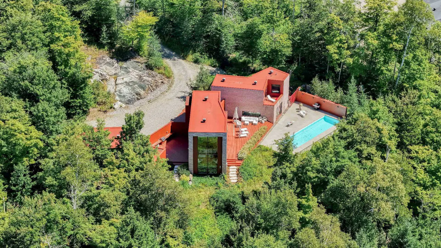Aerial view of a modern house with a red roof, surrounded by green trees, with a pool and patio.