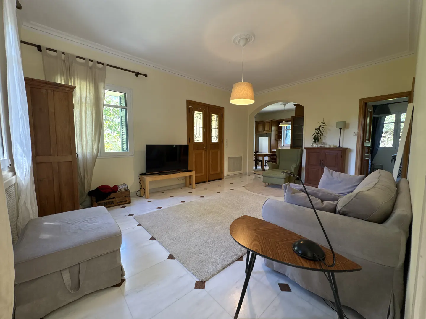 Bright living room with marble floors, a gray sofa, a wooden table, and a TV. A doorway leads to a dining area.