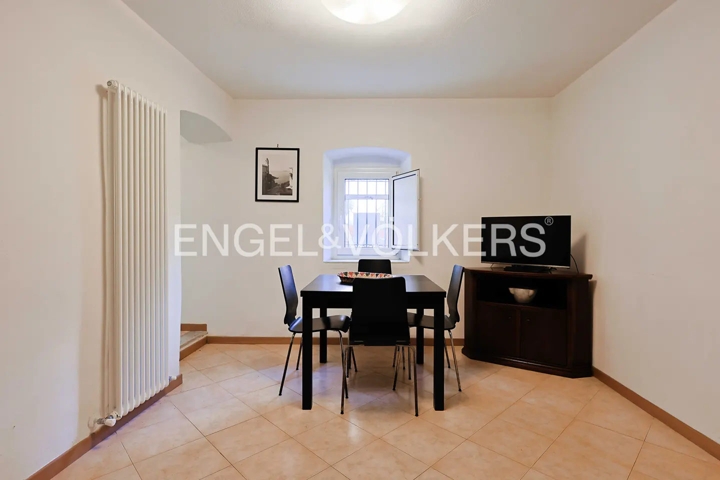 A dining room with a black table and chairs, a TV on a dark wood stand, and a window with white trim. The walls are white, and the floor is tiled.