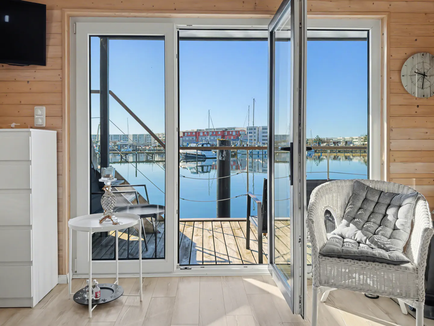 Interior view of a room with wood paneling, open glass doors leading to a marina with boats and buildings in the background.