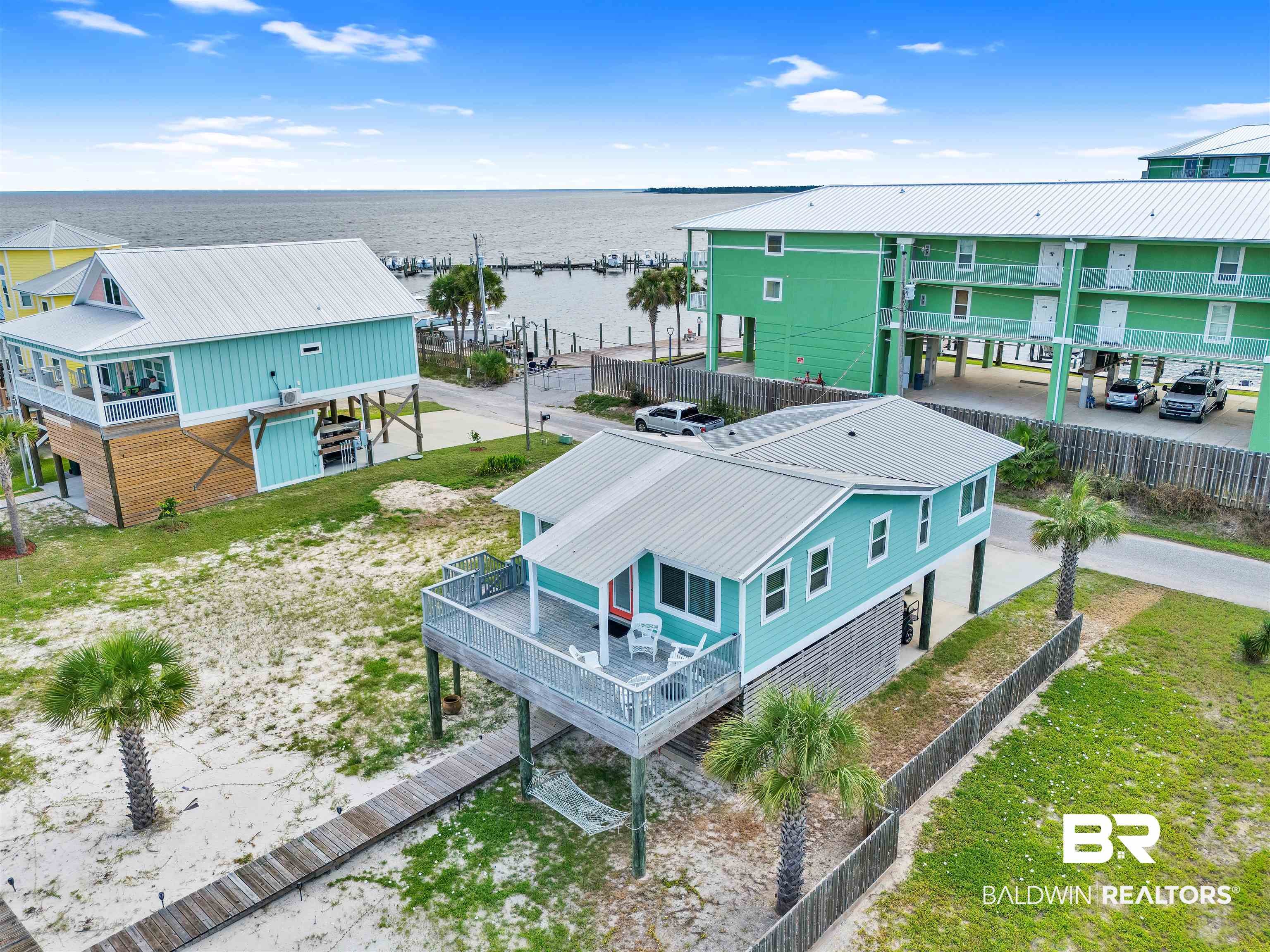 Aerial view of a teal stilt house with a deck, palm trees, and sandy yard near a bay with a pier and green apartments.