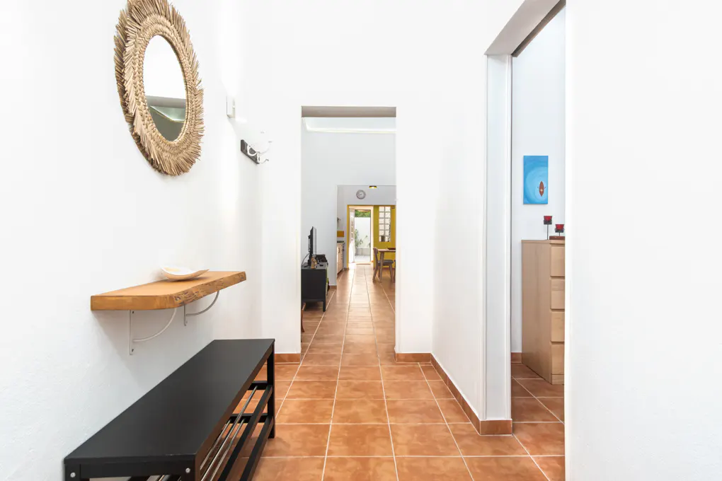 Hallway with white walls and terracotta tile floor. A black bench and wooden shelf are on the left wall, with a straw mirror above. A doorway leads to a dining area.