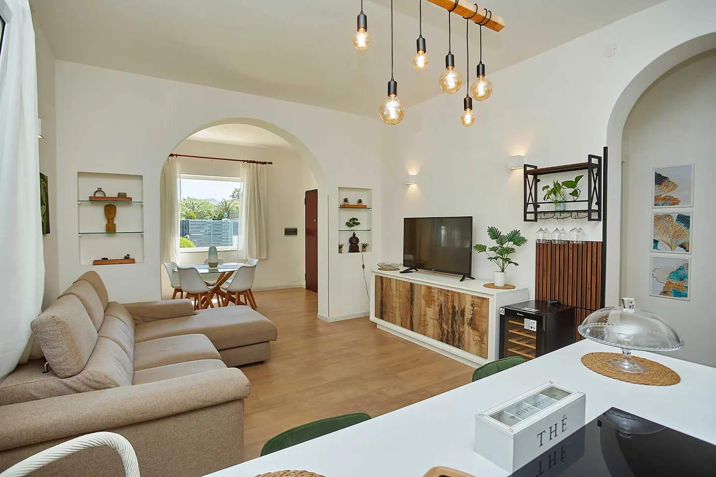 Bright living room with beige sectional sofa, dining table, TV, and modern lighting fixtures in a white-walled home.