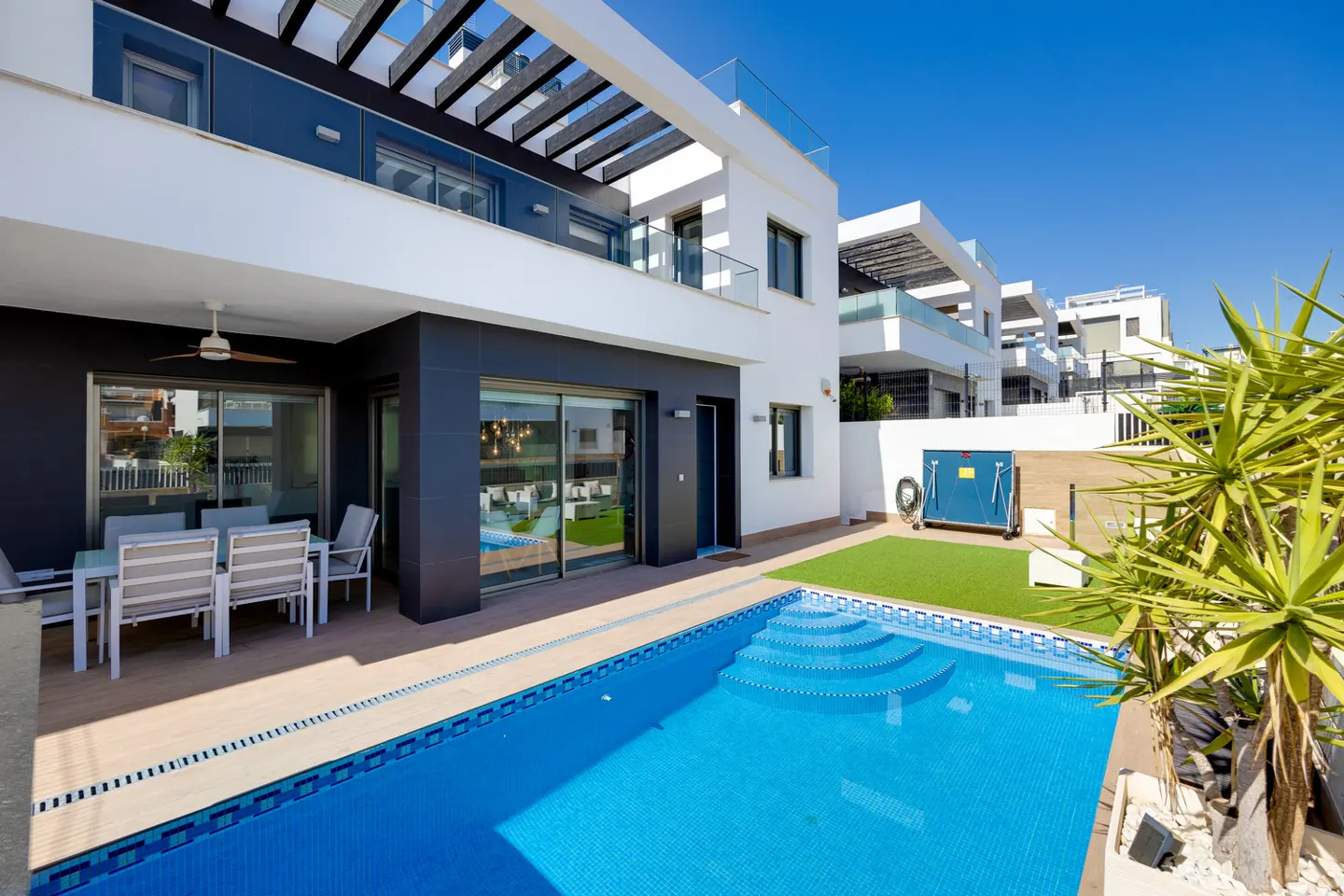 Modern white and dark gray villa with a private swimming pool, patio dining area, and small artificial grass lawn.