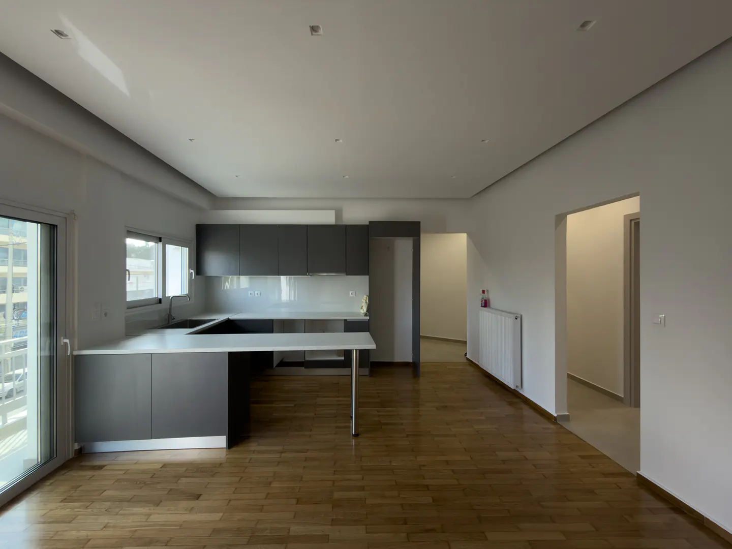 A bright, open-concept apartment with wood floors, a gray kitchen, and white walls. A sliding glass door leads to a balcony.