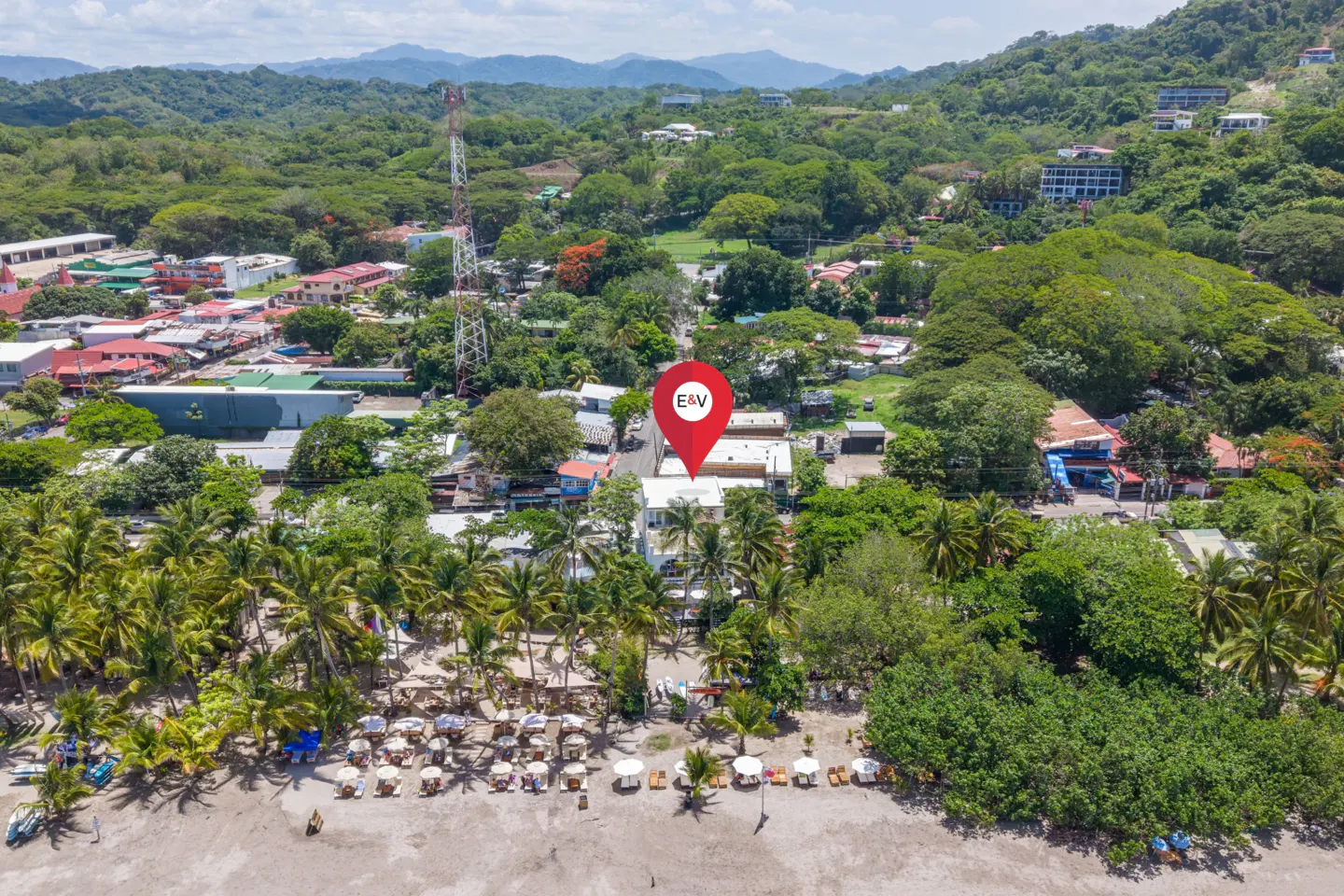 Aerial view of a beach town with a red E&V location pin over a building, surrounded by lush trees and mountains.