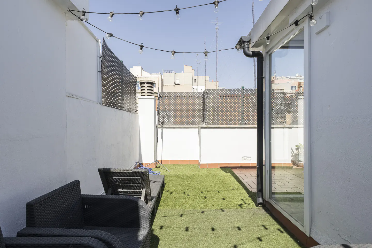 Outdoor patio with artificial grass, black wicker furniture, and string lights against white walls.