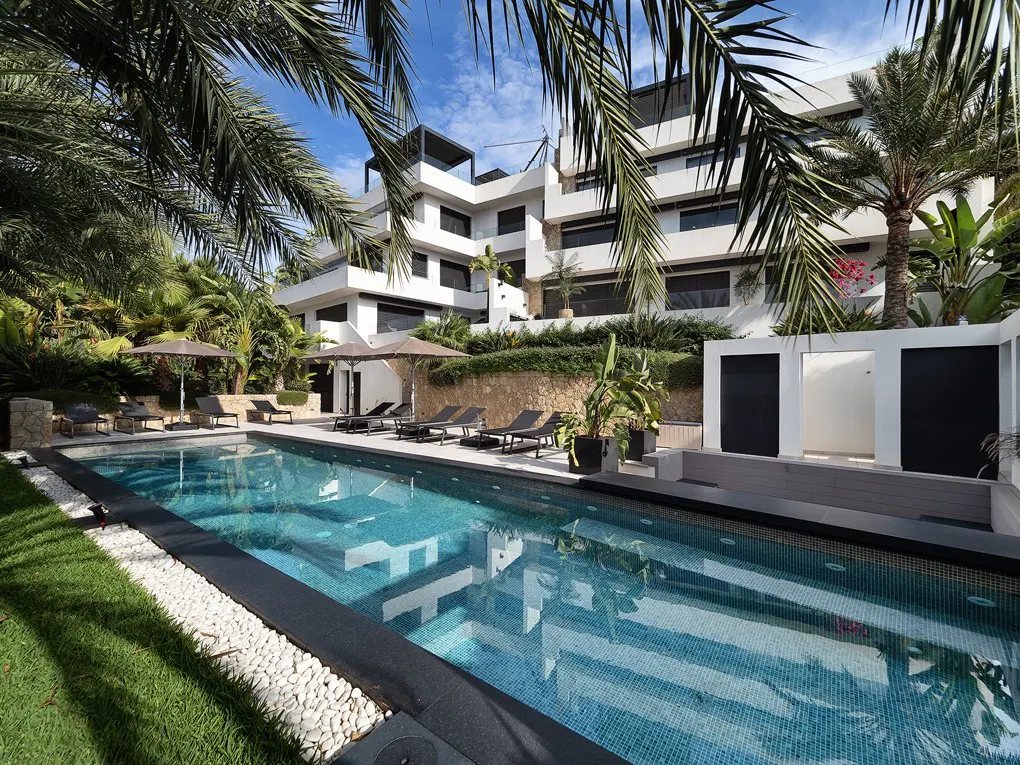 Modern white building with a pool. Lounge chairs and umbrellas surround the pool. Palm trees frame the scene.