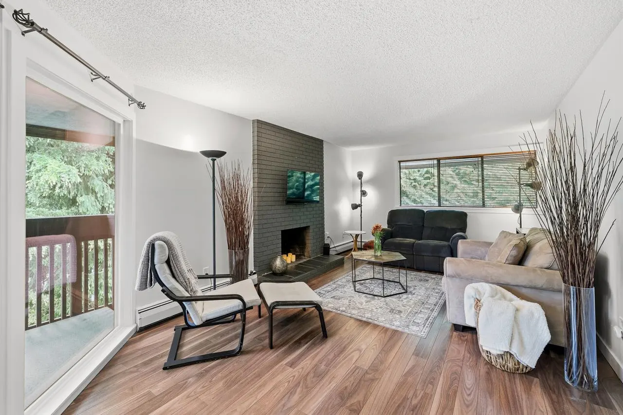 Living room with wood floors, gray brick fireplace, and sofas. A chair faces a balcony with green trees.