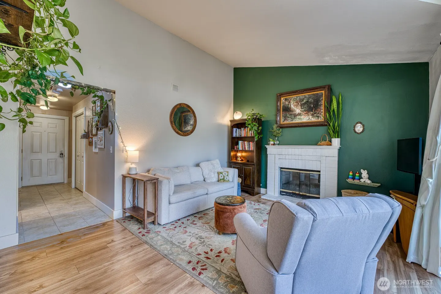 Living room with white sofa, gray armchair, fireplace, and green accent wall. A bookshelf and plants add warmth to the space.