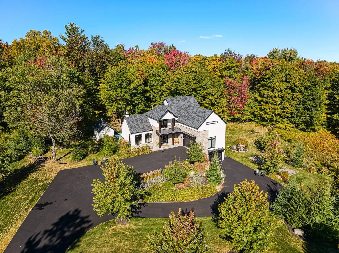 Aerial view of a modern white house with a gray roof, surrounded by colorful autumn trees and a black driveway.