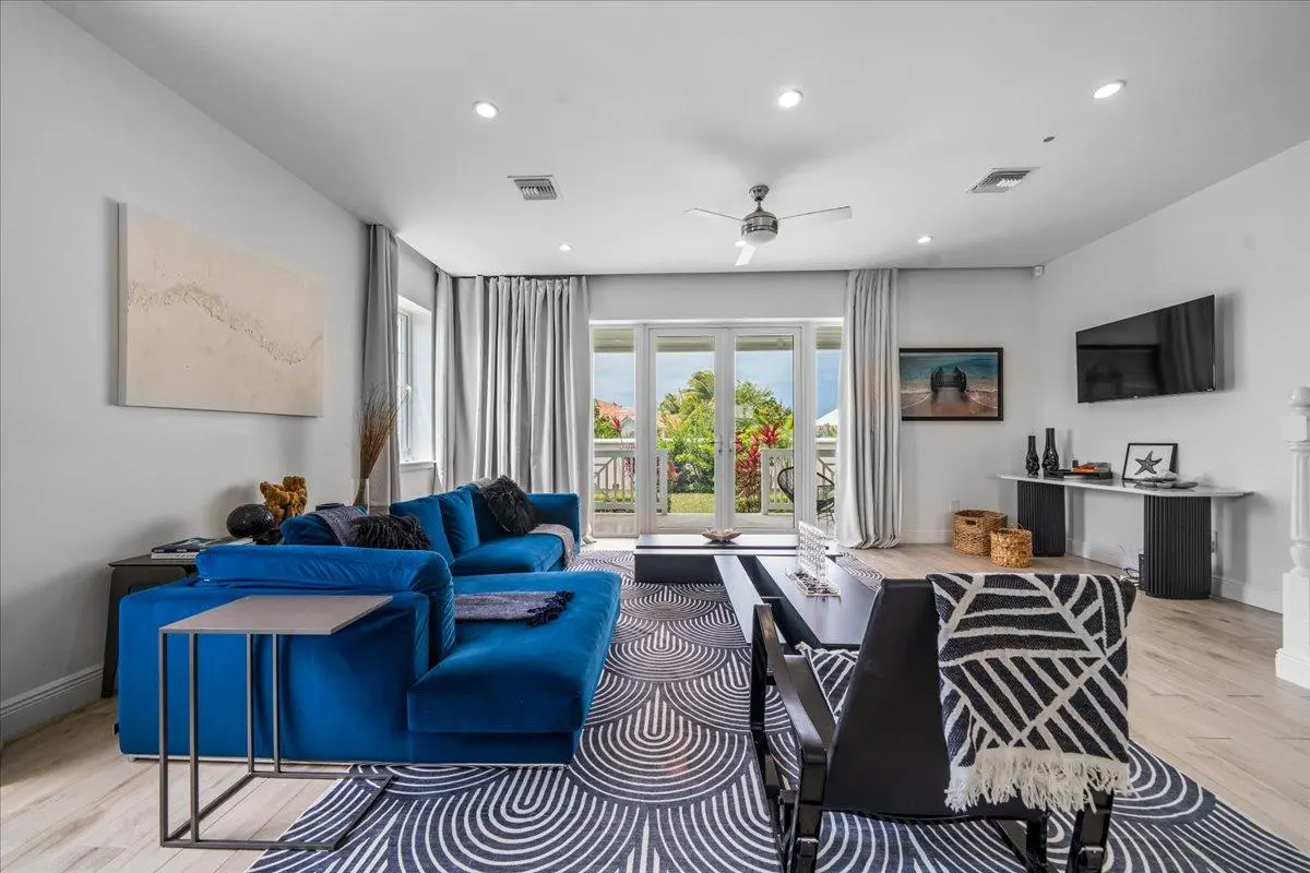Bright living room with a blue velvet sectional sofa, patterned rug, and French doors leading to a balcony with lush greenery.