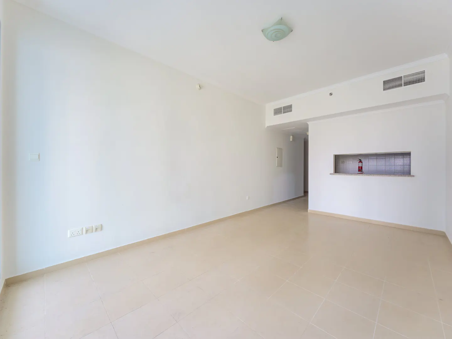 Empty, bright white room with beige tiled floors, a ceiling light, and a small kitchen pass-through window.