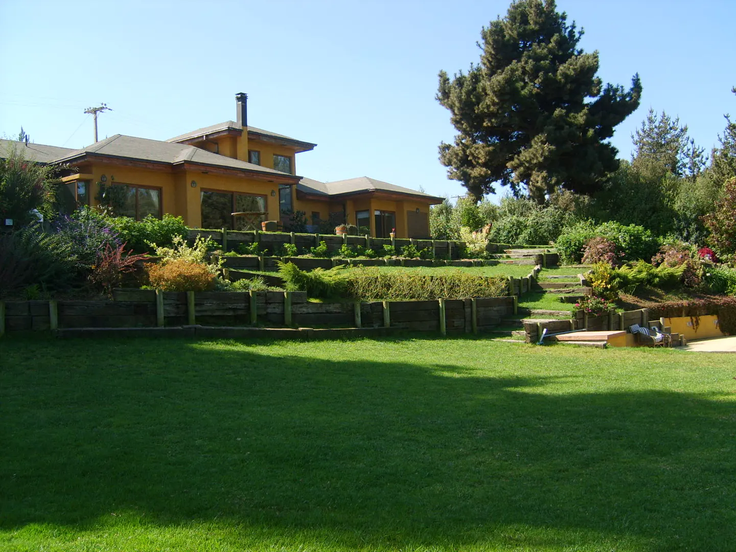 Exterior view of a yellow house with a green lawn and tiered garden under a blue sky.