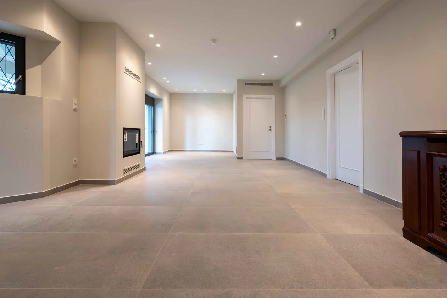 Empty living room with gray tile floor, white walls, recessed lighting, and a modern fireplace.