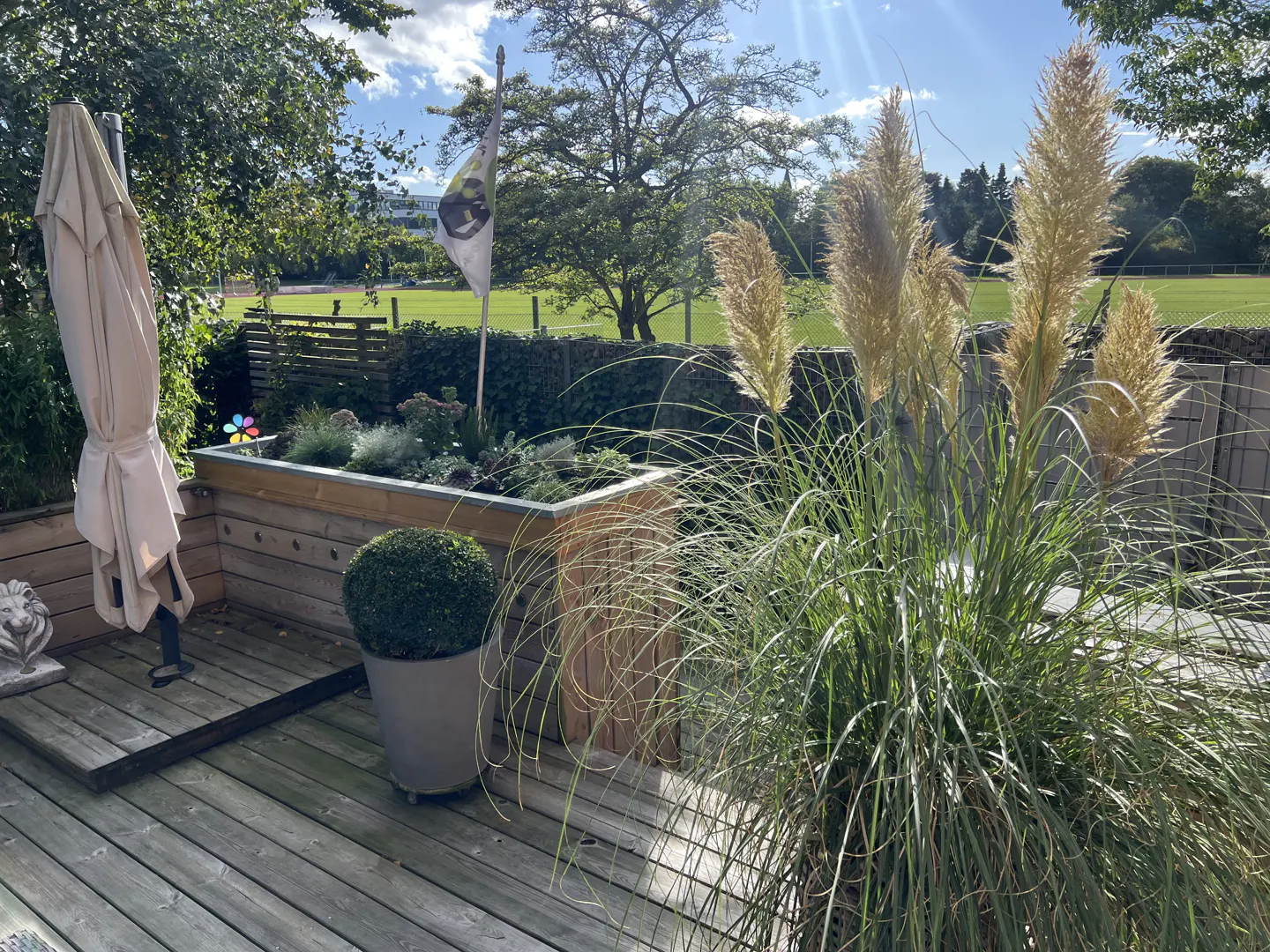 Wooden deck with pampas grass, potted plants, and a closed beige umbrella. A flag and green field are visible in the background.