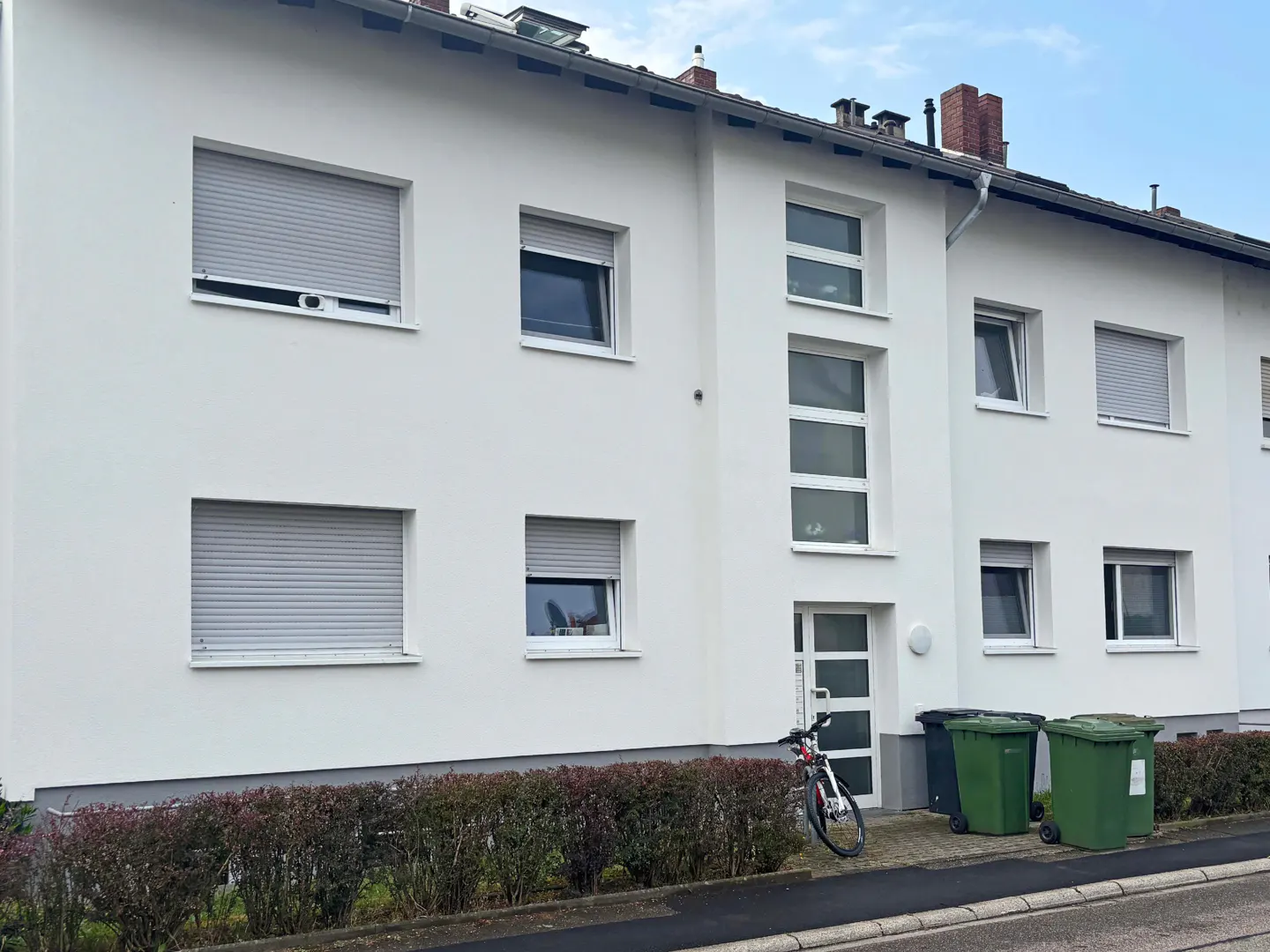 Exterior of a white, two-story apartment building with gray window shutters. A bicycle and green trash bins are near the entrance.