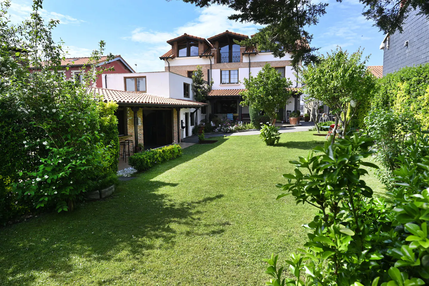 A large green lawn with trees and bushes leads to a two-story house with a red tile roof.