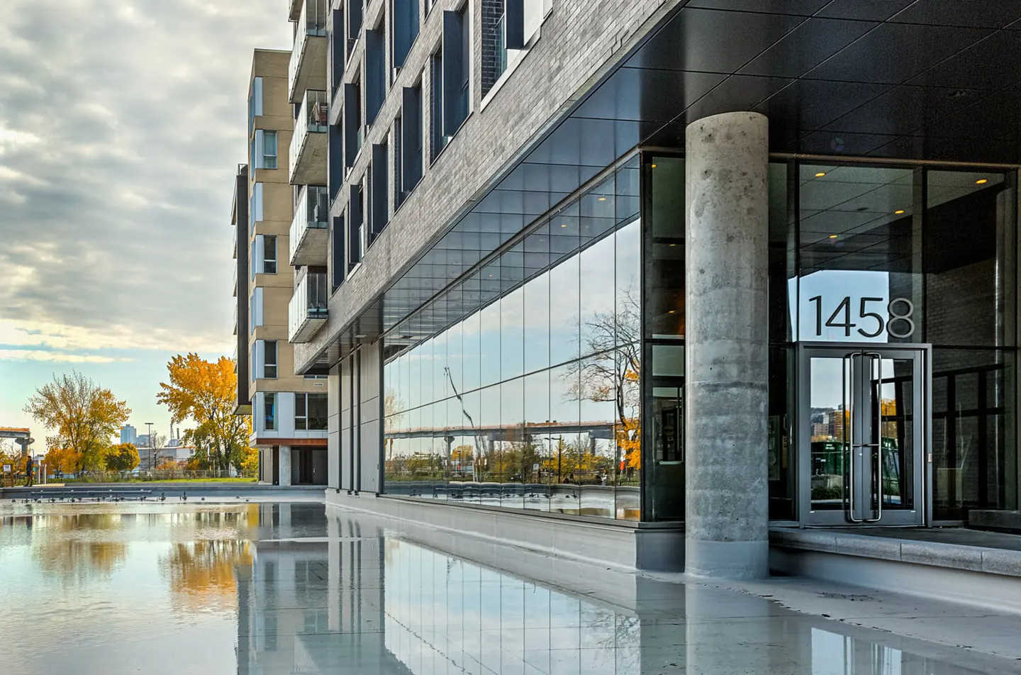 Modern apartment building exterior with glass walls, a concrete pillar, and a reflective water feature at the entrance.