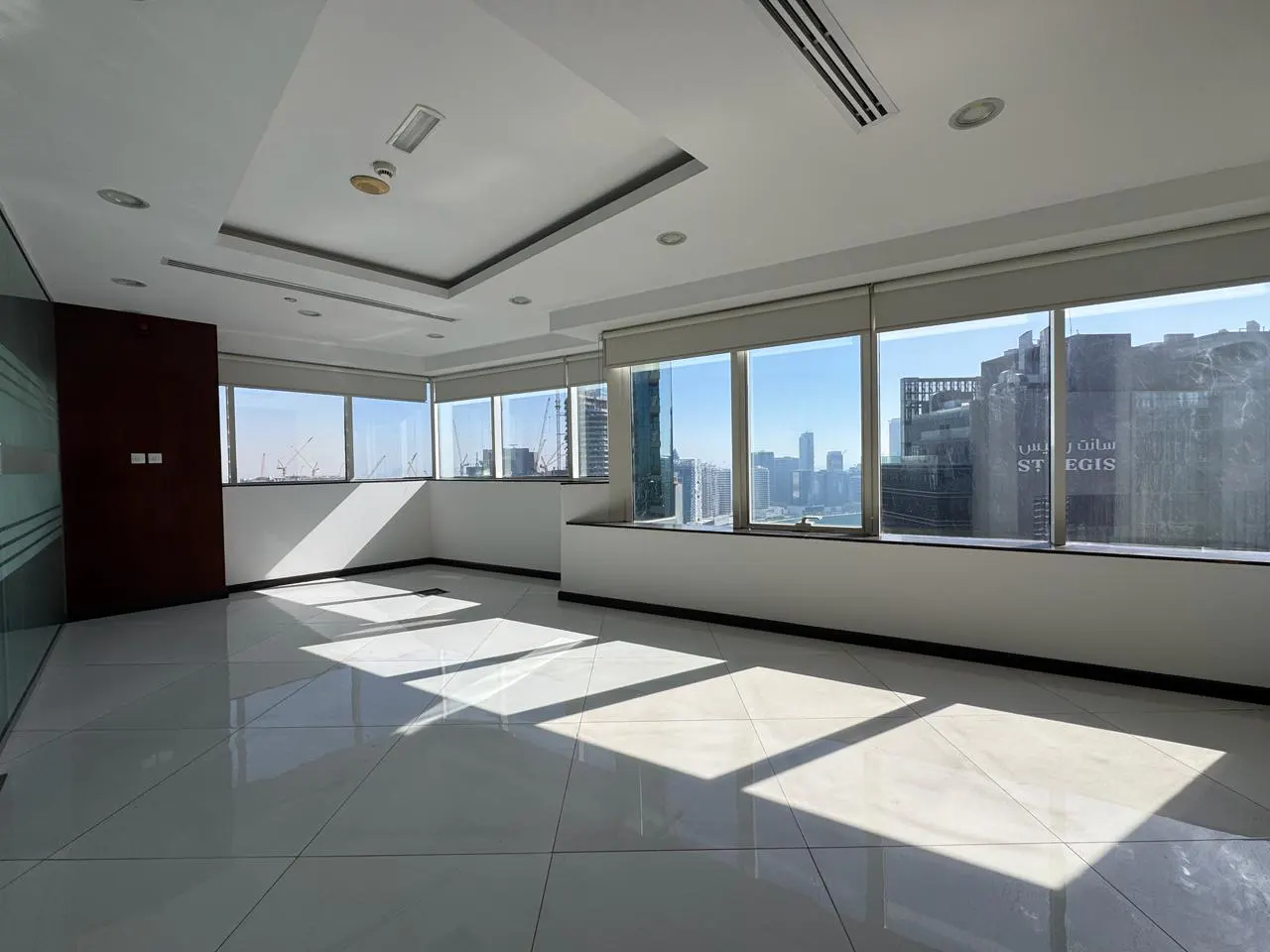 Empty modern office with white tiled floors, recessed lighting, and large windows offering a city skyline view.