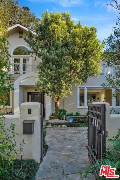 A beige two-story house with a stone walkway, a metal gate, and a large tree in the front yard under a blue sky.