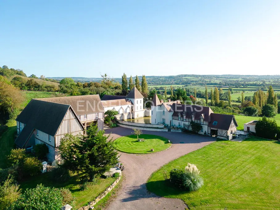 Aerial view of a large, white chateau with a courtyard, surrounded by green lawns and trees under a blue sky.