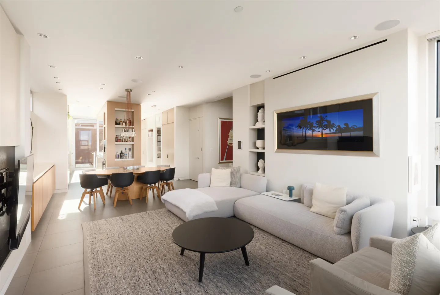 Bright, modern living room with a gray sectional sofa, black coffee table, and dining area with black chairs. A large framed beach scene hangs on the wall.