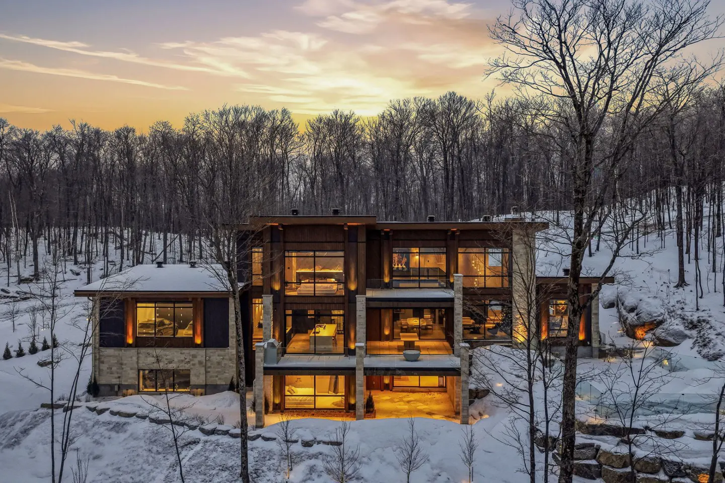 A modern, multi-story home with large windows sits on a snowy hill surrounded by bare trees at sunset.