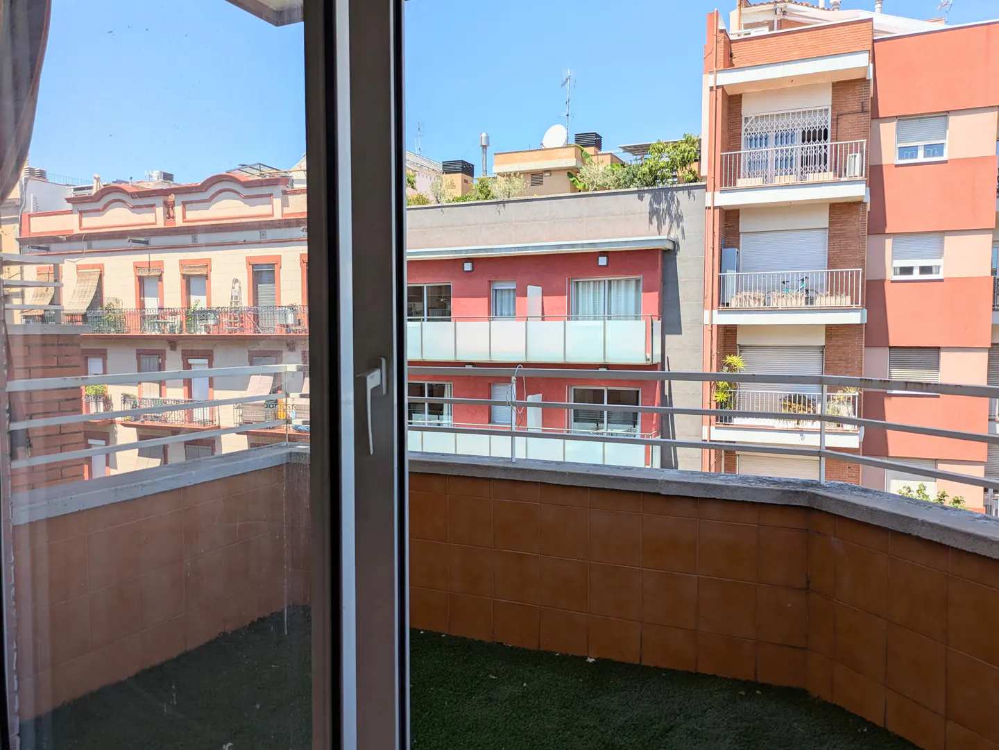 View from a balcony with artificial grass and tiled walls, overlooking neighboring apartment buildings on a sunny day.