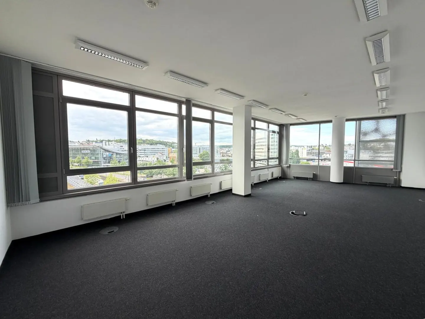 Bright, empty office space with black carpet, white walls, and large windows overlooking a city.