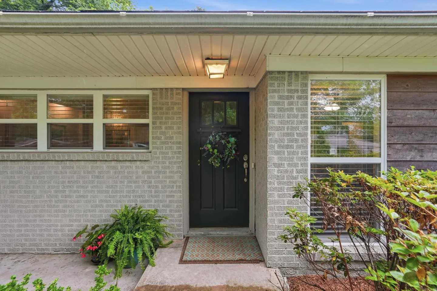 Front view of a one-story house with a black door, brick facade, and green wreath. Windows with blinds are on either side of the door.