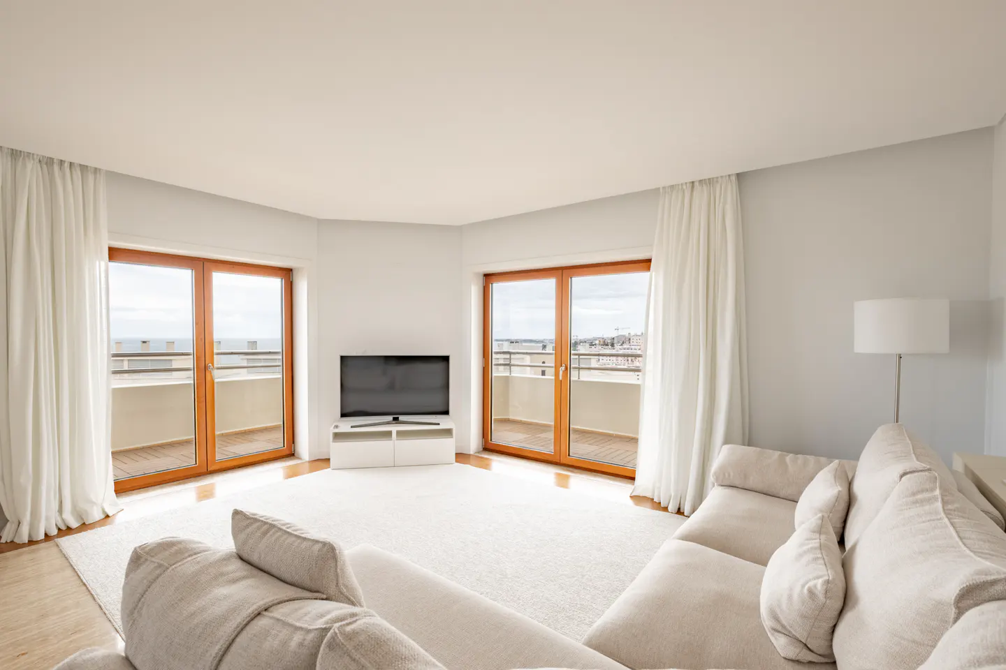 Bright living room with white walls, sofa, and curtains. Wood-framed glass doors lead to balconies with ocean views. A TV sits on a white stand.