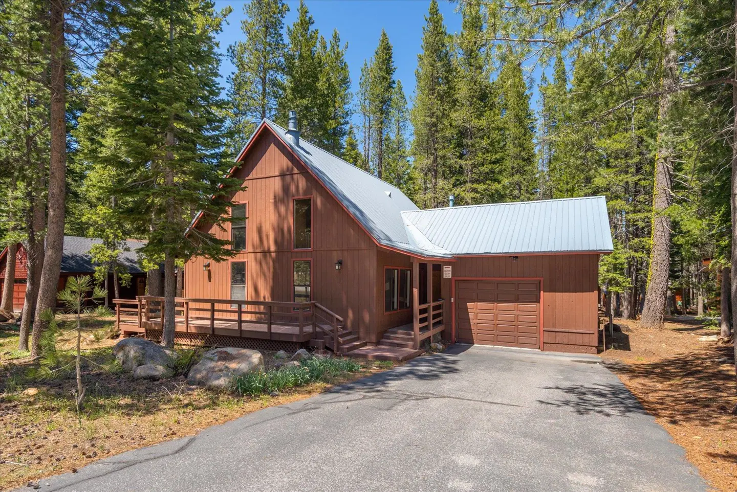 A brown, two-story house with a metal roof, surrounded by tall green trees and a paved driveway.