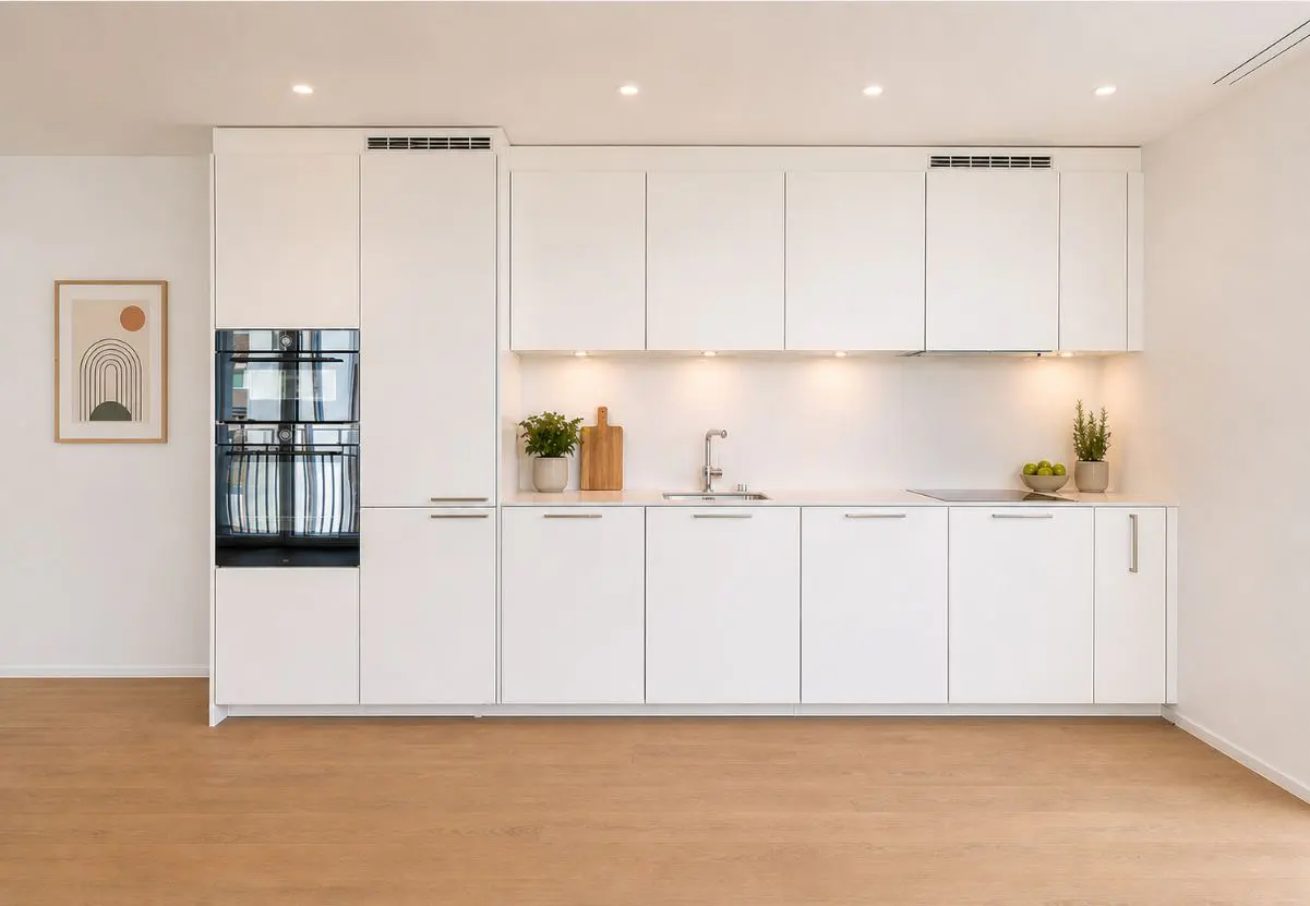 Bright, modern kitchen with white cabinets, stainless steel sink, and wood floors. An oven is built into the cabinets. A framed print hangs on the wall.
