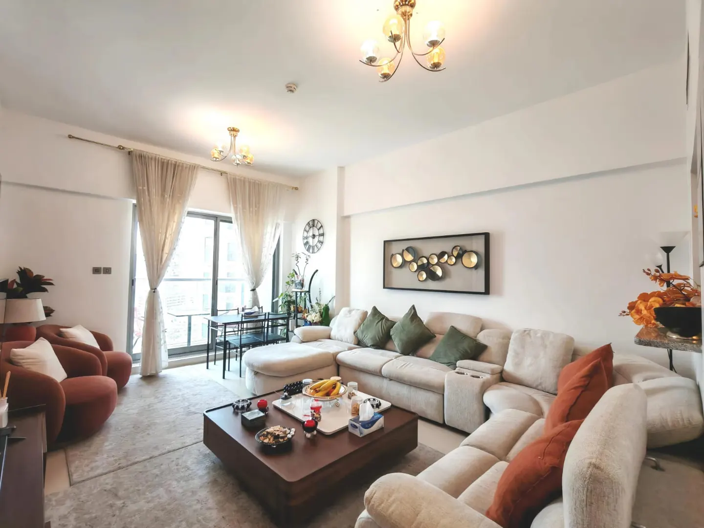 Bright living room with a beige sectional sofa, brown coffee table, and two rust-colored armchairs. A dining table sits by the window.