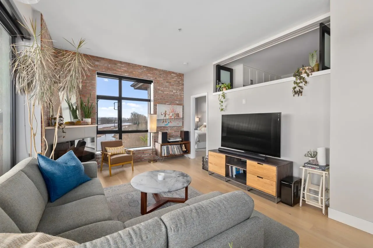 A modern living room with a gray sofa, brick wall, large window, and a loft area above the TV.