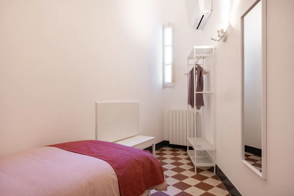 A small bedroom with a bed, bench, white rack with a scarf, and a mirror. The floor is tiled in a checkered pattern.