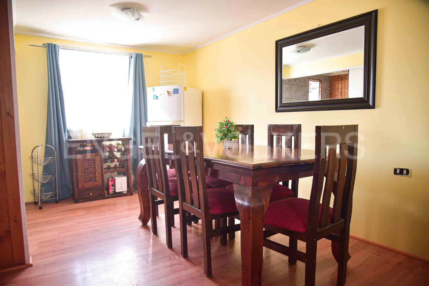Dining room with yellow walls, wooden table and chairs with red cushions, a mirror, and a wine rack near a window.