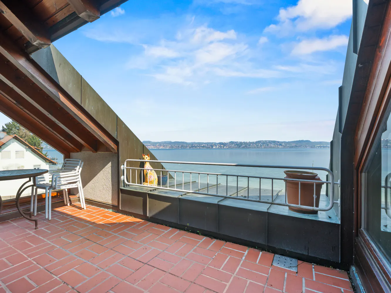 A brick-paved balcony with a table, chair, and a railing overlooking a lake under a blue sky.