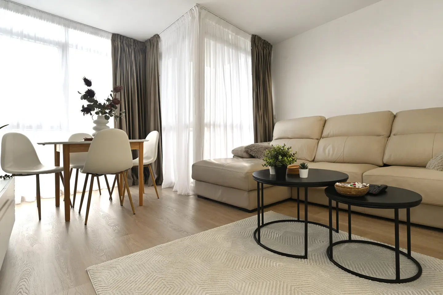 Bright living room with a beige sectional sofa, black oval coffee tables, and a dining table with white chairs. Large windows with sheer and brown curtains.