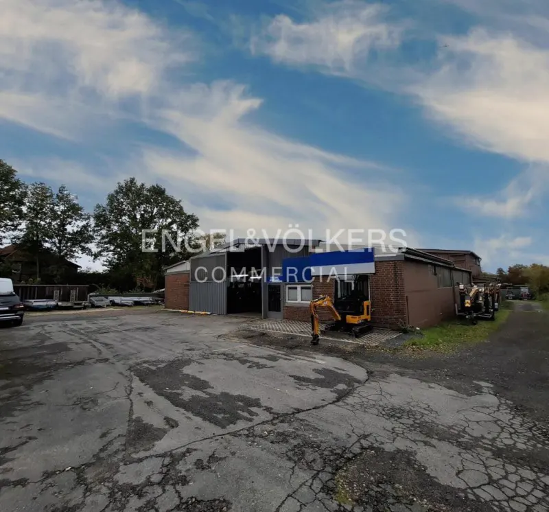 Exterior of a one-story commercial building with Engel & Völkers signage, a small excavator, and a cracked asphalt parking lot.