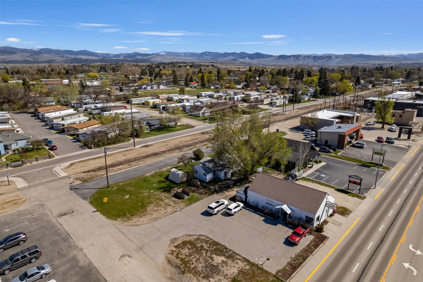Aerial view of a small town with houses, businesses, a railroad track, and mountains in the background under a clear blue sky.