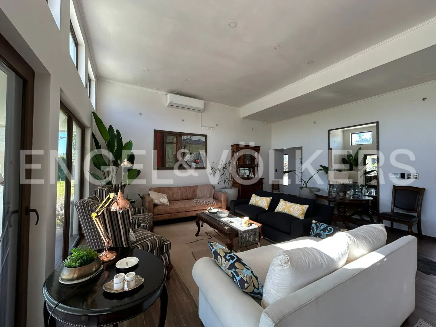 Bright living room with white walls, large windows, and multiple sofas. A painting hangs above a brown sofa. A black table sits in the foreground.