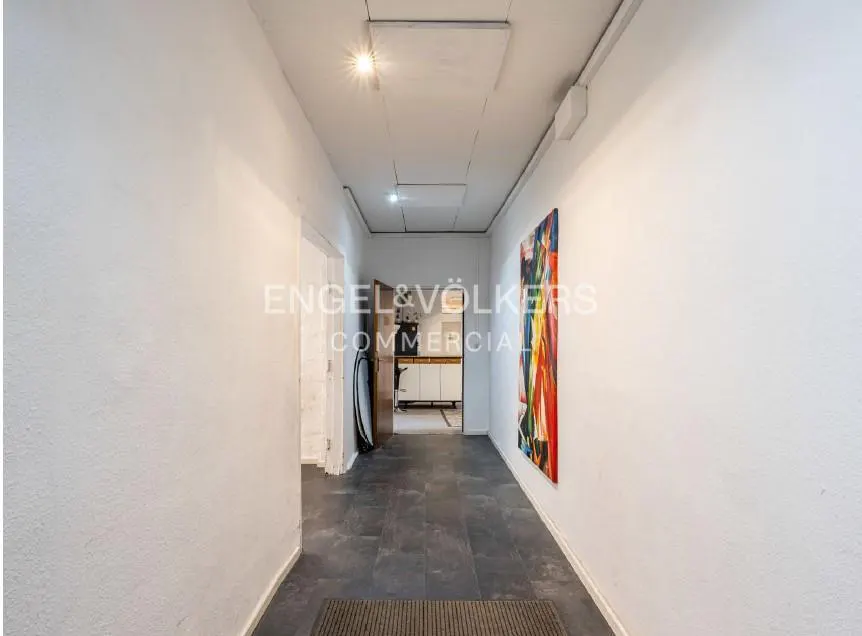 Hallway with white walls, gray tile floor, and a colorful painting on the right wall. A doorway leads to a kitchen in the background.