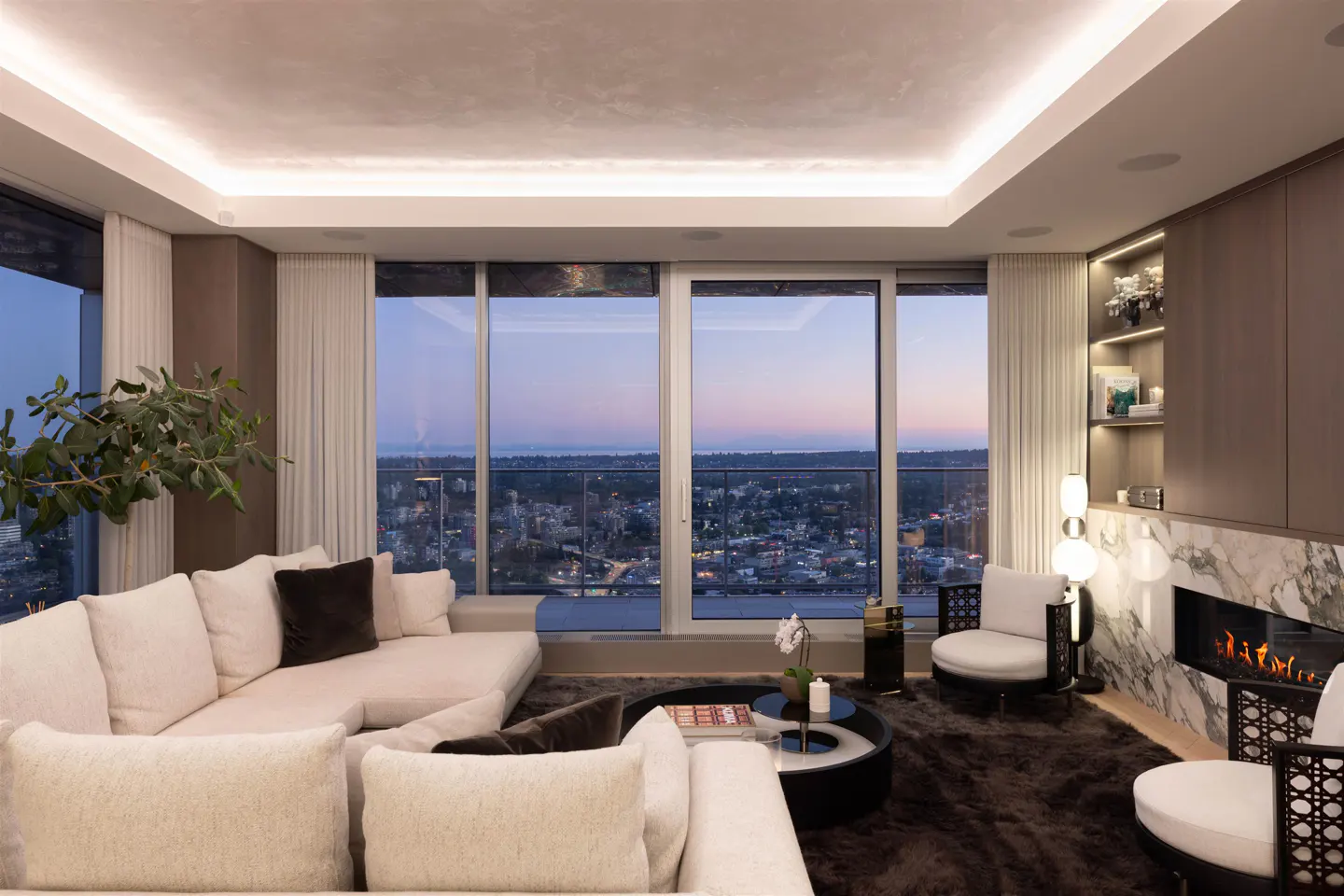A modern living room with a white sofa, chairs, and a fireplace. Large windows offer a city view at dusk.