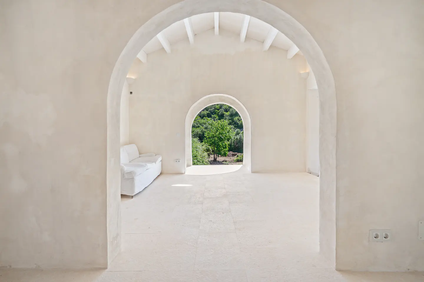 Minimalist white room with arched doorways, a white sofa, and a view of lush green trees through the back archway.