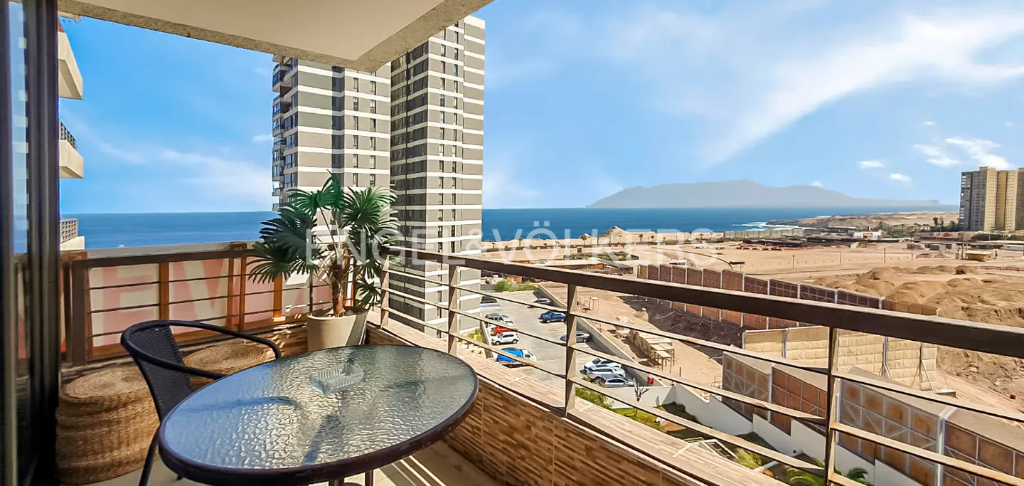 Balcony view with a glass table, chair, and potted plant overlooking the ocean, a city, and a mountain under a blue sky.